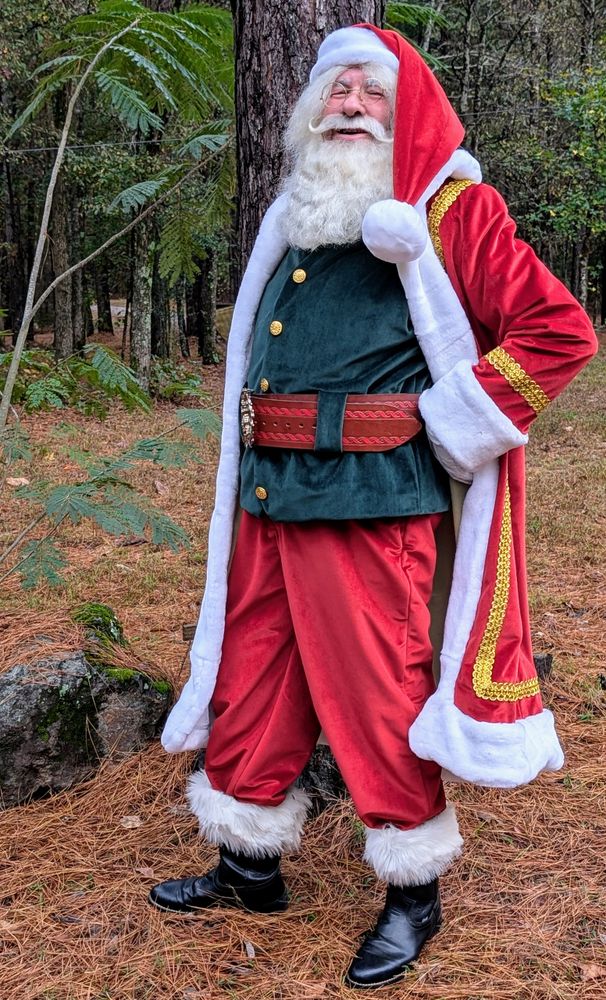 Santa Claus in a festive red and green outfit stands smiling in a forest setting.