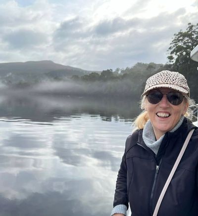 Smiling woman in sunglasses and hat by a misty lake with mountains in the background.