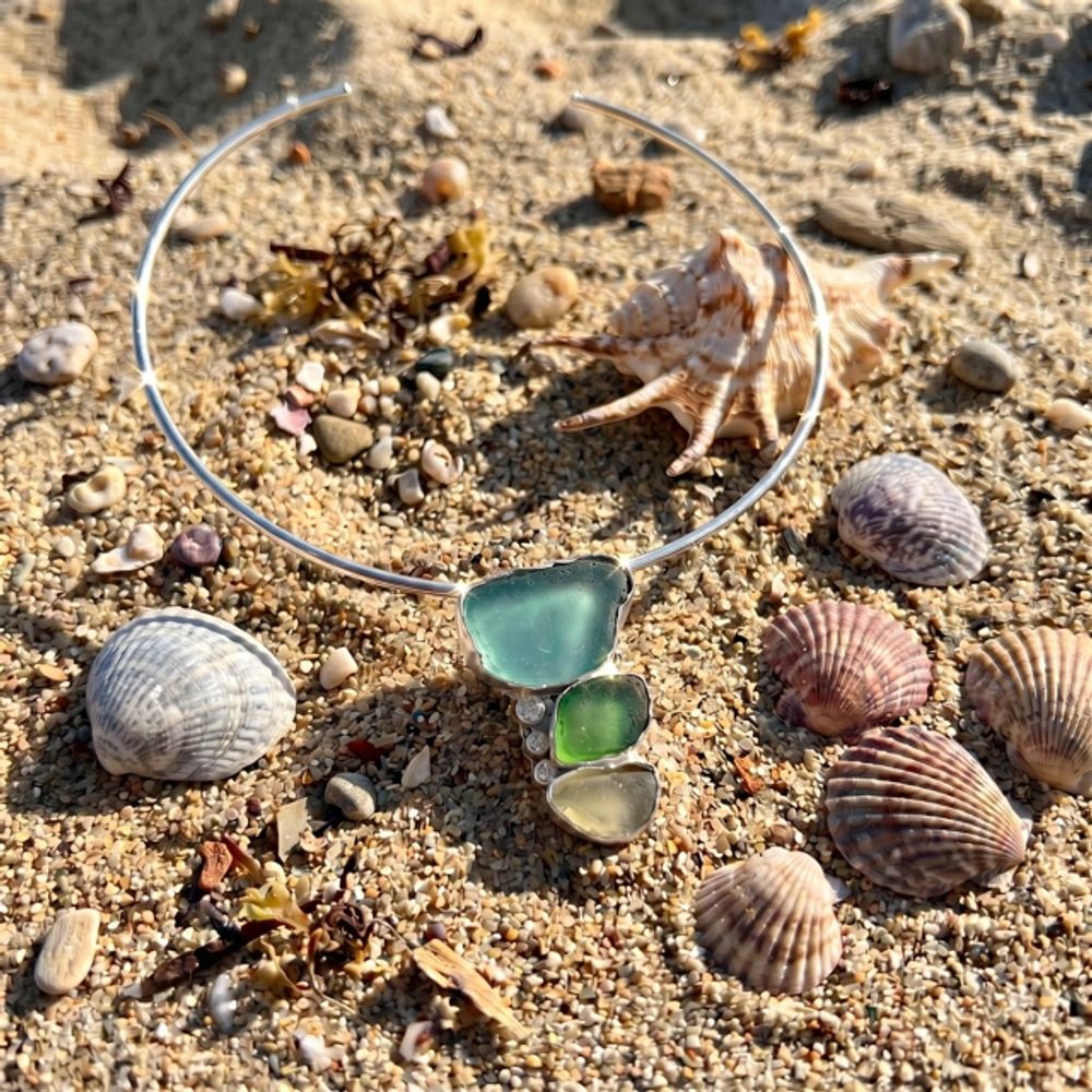 Silver coker necklace with sea glass and shells on sandy beach.
