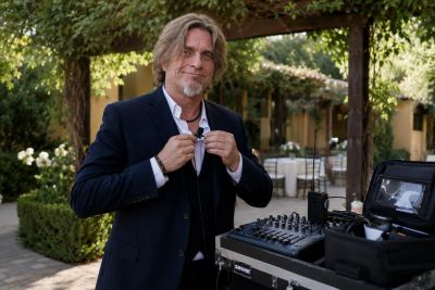 Man in a suit adjusting a microphone near audio equipment outdoors.