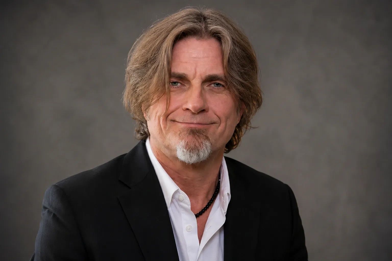 Man with medium-length hair and a goatee, wearing a black suit jacket and white shirt.
