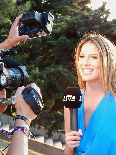 A blonde woman in blue interviewed outdoors with a camera and microphone.