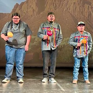 Three men in traditional attire holding rattles on stage.