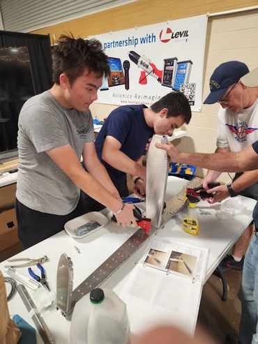 Three people assembling a metal component at a workshop table.