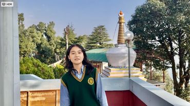 A young girl in a school uniform stands on a balcony with green trees and a stupa in the background.