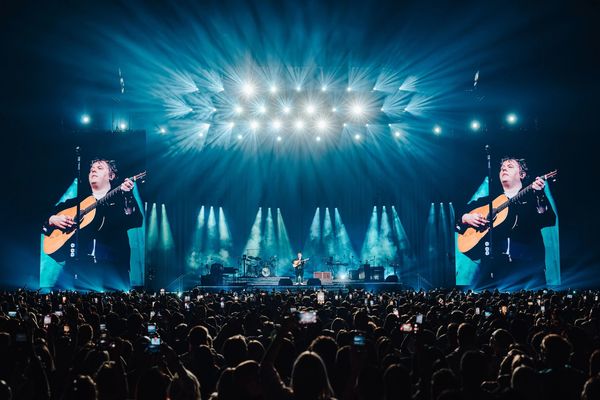 Wide angle shot of Lewis Capaldi playing guitar on stage. Beams of light shoot in to the audience.