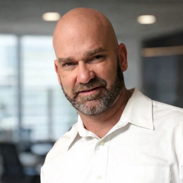 A bald man with a beard wearing a white shirt, smiling slightly indoors.