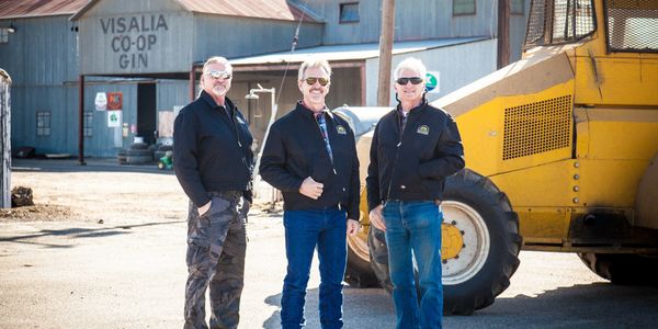 Three men standing outdoors near a yellow vehicle and a coop building.