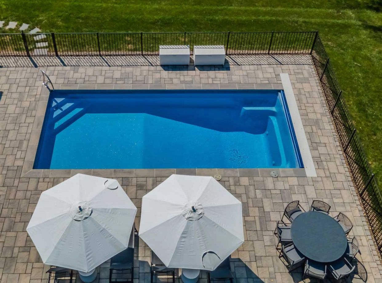 A rectangular swimming pool with white umbrellas and outdoor seating on a tiled deck.