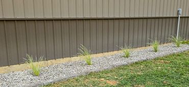 Strip of gravel with grass plants along a metal fence.