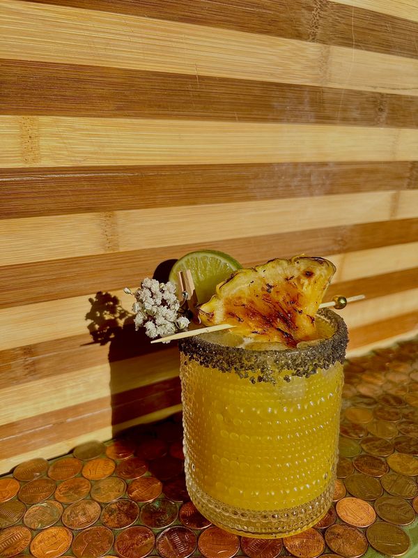 A tropical cocktail garnished with grilled pineapple, lime, and flowers on a penny-covered surface.