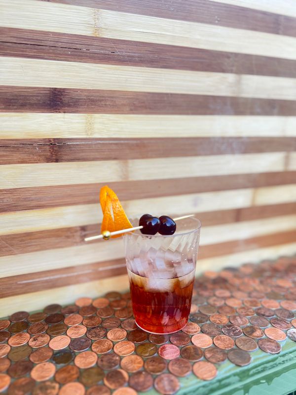Cocktail with cherries and orange peel on a penny-covered surface.