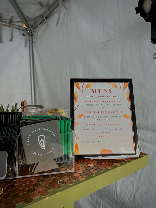 A drink menu and branded koozies on a penny-covered table at an event.