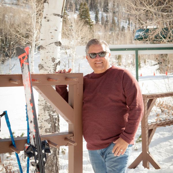 Man in sunglasses and maroon sweater standing by skis in snowy outdoors.