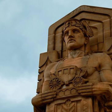 A monumental stone statue of a man holding a covered wagon against a cloudy sky.