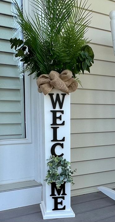 Tall white planter with greenery and 'WELCOME' letters on a porch.