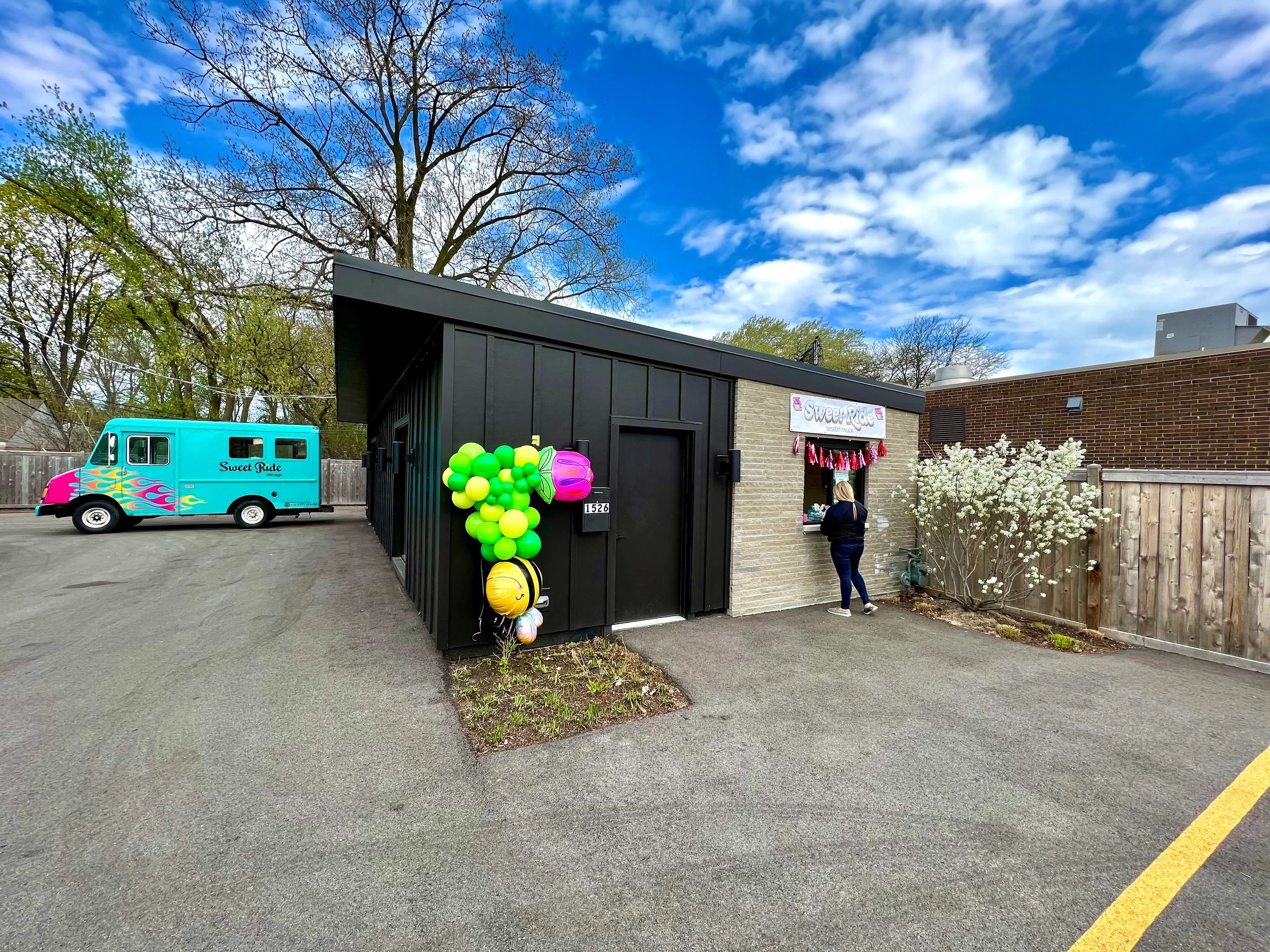 stand alone building with balloons and a walk-up window with a teal food truck parked in back