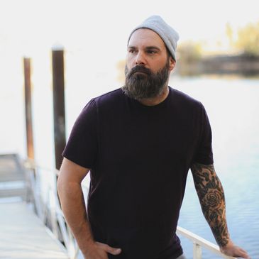 Bearded man with a tattooed arm wearing a beanie and dark t-shirt by the water.