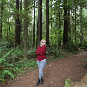 Woman in red plaid shirt standing on forest path surrounded by tall trees.