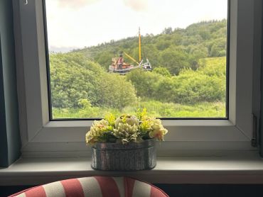 Flower pot on a windowsill with a boat and green hills outside.