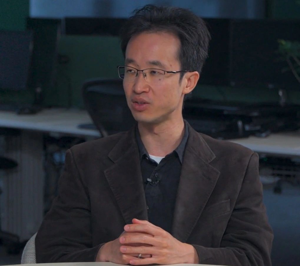 Man in glasses and dark blazer sitting at a table speaking.