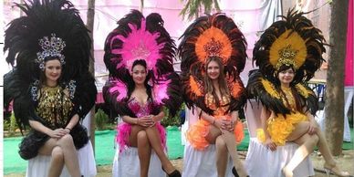 Four women in colorful feathered costumes sitting on chairs outdoors.
