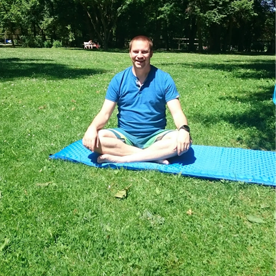 Man sitting cross-legged on a blue mat in a sunny park.