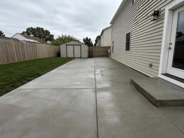 A clean concrete patio beside a house with a small shed and wooden fence.