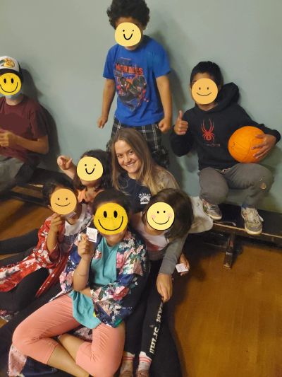 A group of children and a woman posing happily indoors, one child holding a pumpkin.