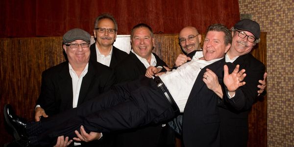 Six men in suits, five holding another man horizontally, all smiling joyfully.