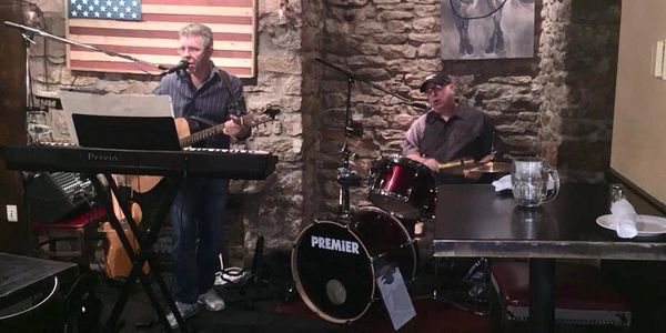 Two musicians performing live in a cozy venue with stone walls and an American flag.