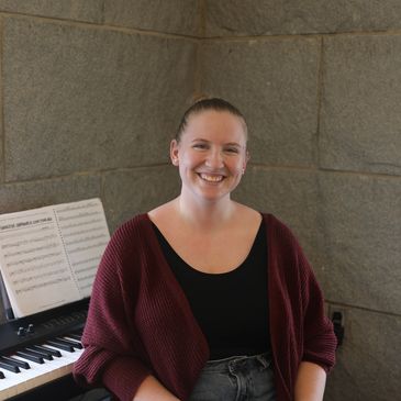 Smiling woman sitting by a keyboard with sheet music in the background.