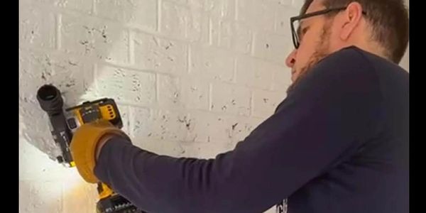 Man using a drill on a white brick wall wearing safety gloves and glasses.