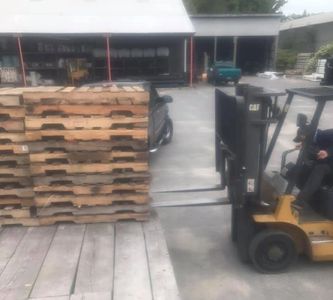 A forklift driver unloading a stack of wood pallets off of the back of a flatbed truck