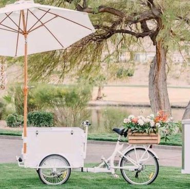 White tricycle cart with umbrella and flower box in a park.