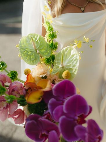 Bride holding a vibrant bouquet with orchids and anthuriums in soft sunlight.