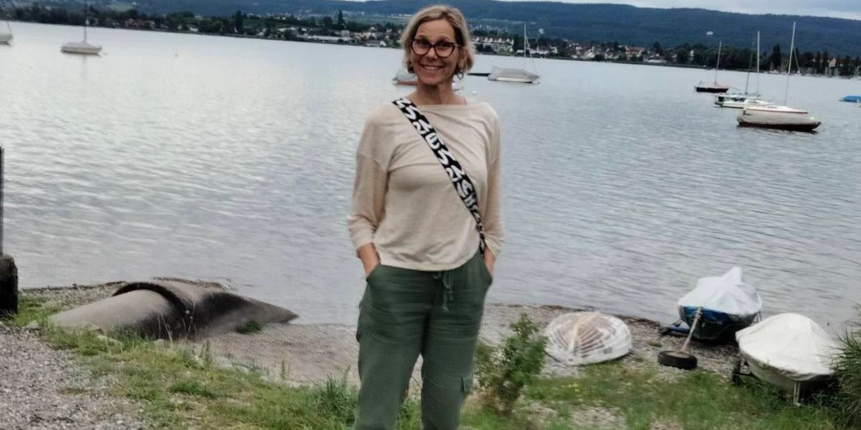 A smiling woman stands by a lakeside with boats and hills in the background.