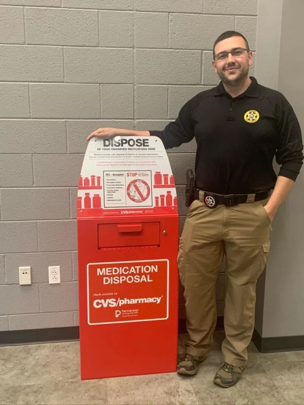 Man in uniform stands next to a red medication disposal box at CVS Pharmacy.