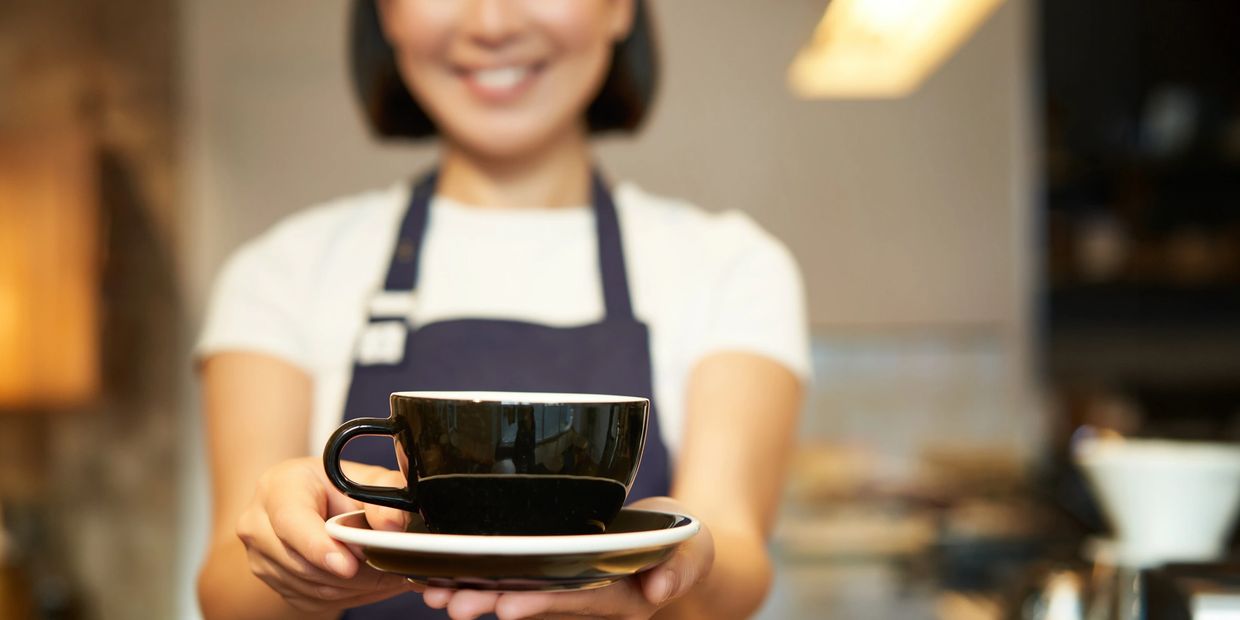 Smiling barista offering a black coffee cup on a saucer.