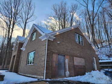 Brick house with snow-covered roof and boarded-up door in winter woods.