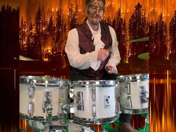 A masked drummer plays in front of a forest fire backdrop.