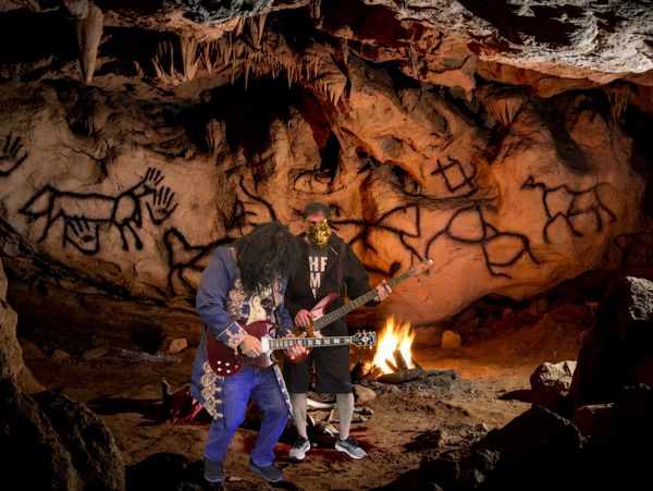 Two musicians play guitars in a cave with prehistoric wall art and a campfire.