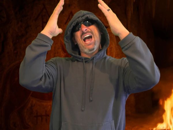 Man in hoodie and sunglasses passionately yelling with hands raised.