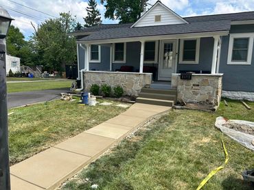 Newly poured concrete sidewalk leads to a quaint house with a stone porch.