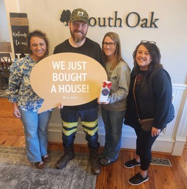 Four people celebrate buying a house with a large sign indoors.