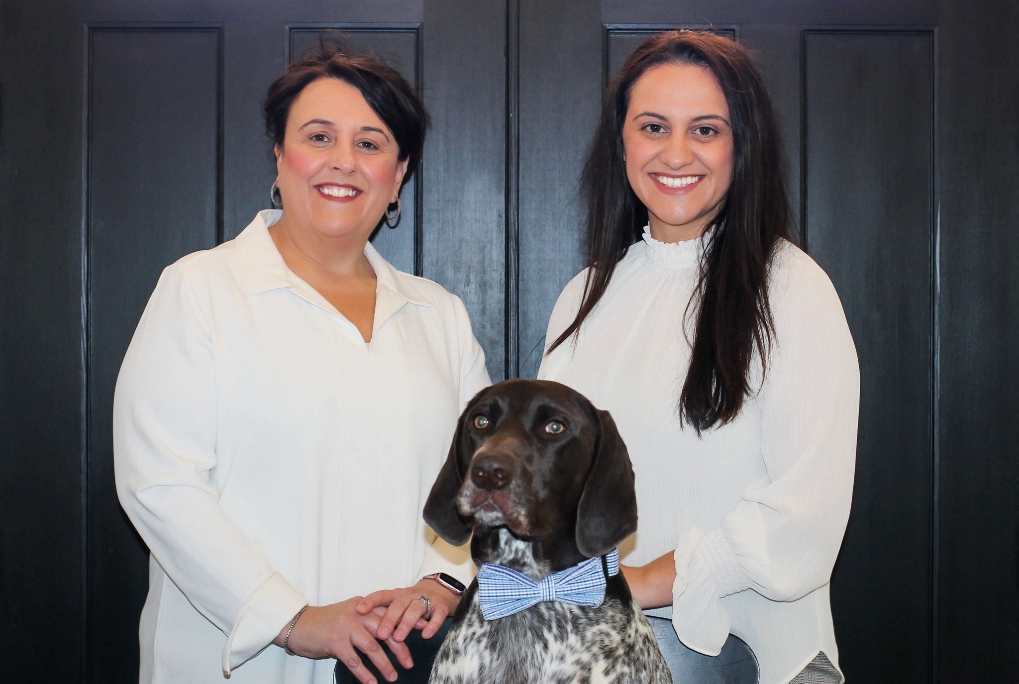 Two women smiling with a dog wearing a blue bow tie.
