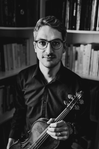 Man with glasses holding a violin in a library setting.
