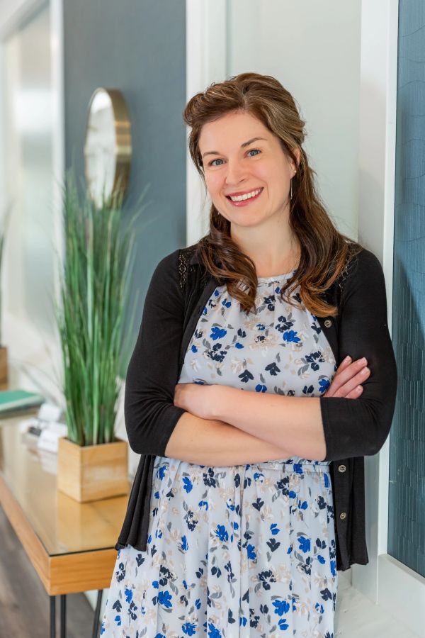 Smiling woman in floral dress and black cardigan stands with arms crossed indoors.