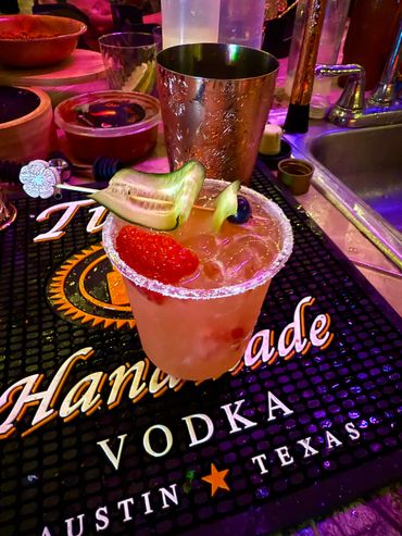 Colorful cocktail garnished with cucumber, strawberry, and blueberry on a bar mat.