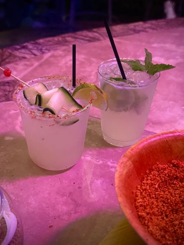 Two refreshing cocktails with cucumber, lime, and mint garnishes on a pink-lit table.